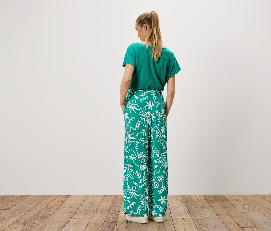 Eine Frau steht mit dem Rücken zur Kamera, sie trägt ein grünes T-Shirt und eine grüne Hose mit weißem Muster.