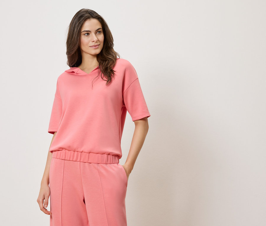 Frau trägt einen pinkfarbenen Kapuzenpullover mit kurzen Ärmeln und eine pinkfarbene Hose.