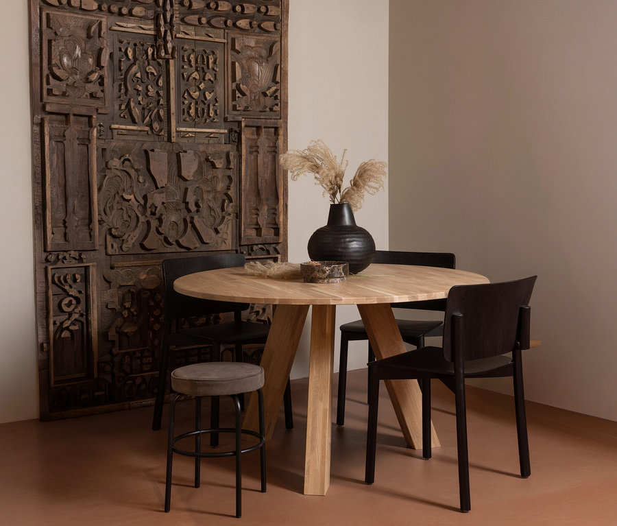 Ein runder, heller WOOOD »Rhonda« Esstisch, ca. 129 cm, steht mit vier Stühlen und einem Hocker vor einer verzierten Holzwand.