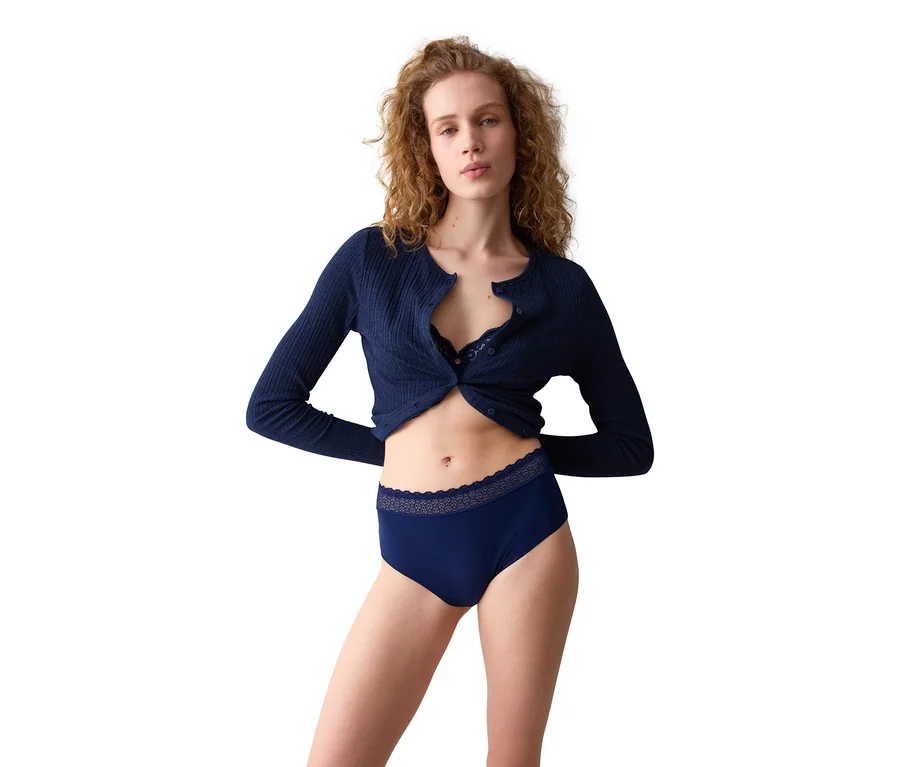 Eine Frau mit lockigem Haar trägt einen marineblauen gerippten Cardigan, einen marineblauen BH und marineblaue Slips mit Spitzenbesatz, während sie in die Kamera blickt.