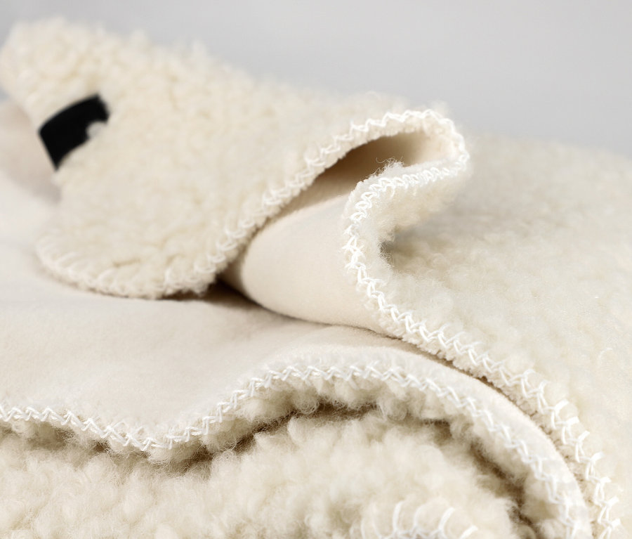 Nahaufnahme einer cremefarbenen, flauschigen Decke, die gefaltet ist, um ihre weiche, wollige Textur und eine glattere Unterseite zu zeigen. Weiße Nähte sind an den Rändern sichtbar, und ein kleines schwarzes Etikett ist in eine Falte gesteckt.