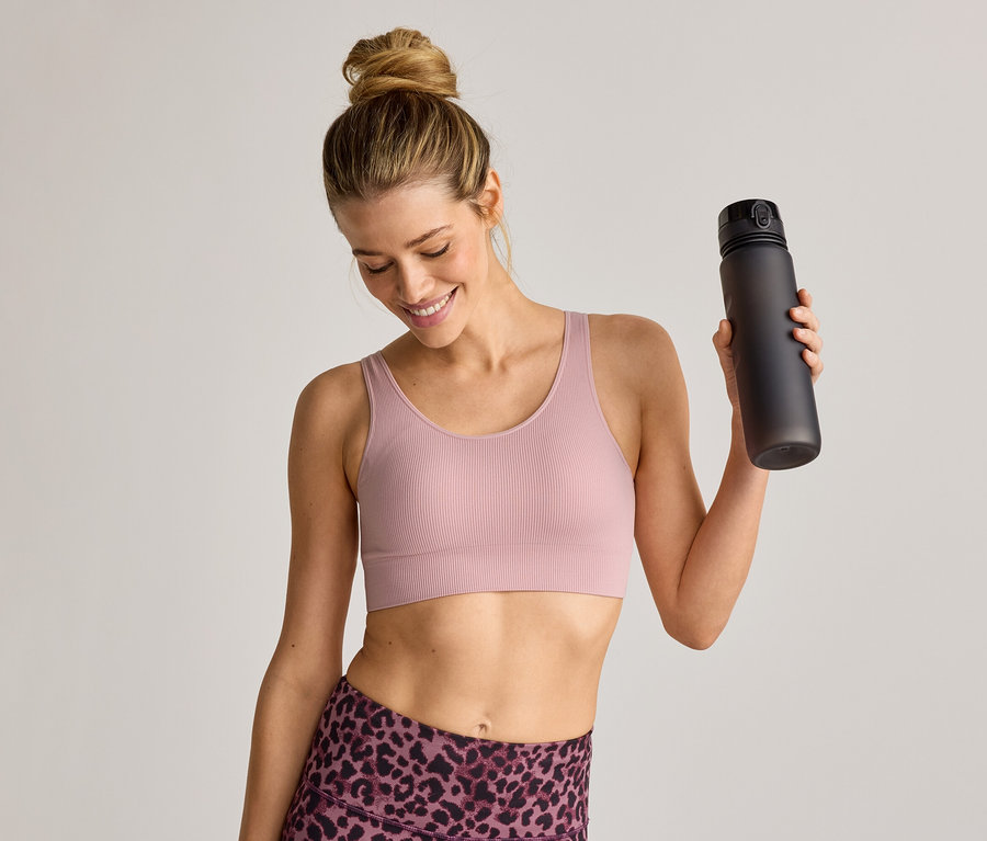 Frau im Sport-BH und Leggings hält eine schwarze Wasserflasche und lächelt.