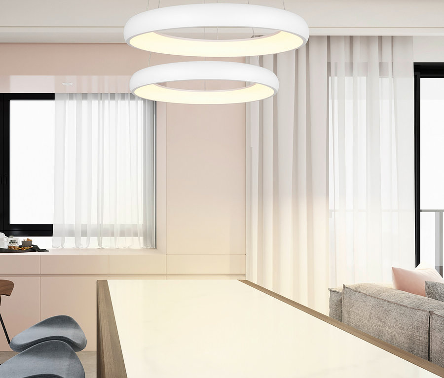 Zwei weiße, ringförmige Pendelleuchten hängen über einem langen Esstisch mit heller Platte und Holzrahmen. Graue Stühle stehen am Tisch, im Hintergrund sind Fenster mit weißen Vorhängen und ein graues Sofa mit rosa Kissen zu sehen.