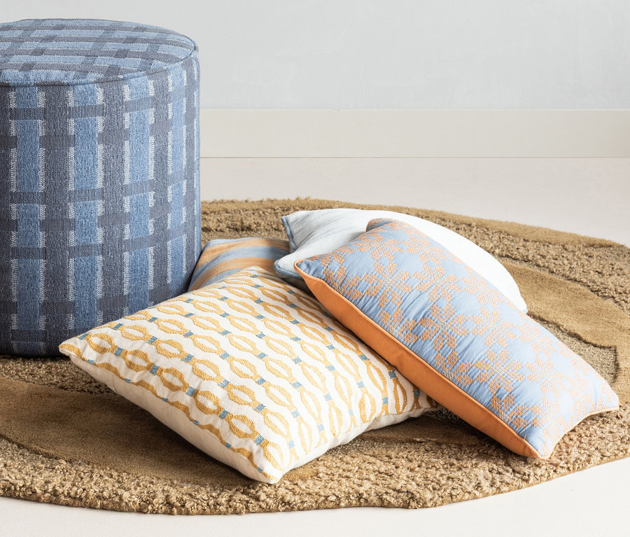 Runder WOOOD Teppich »Orvi«, Ø 160 cm, mit blauem Hocker und drei Zierkissen. Eines weiß mit gelb-blauem Kettenmuster, ein weiteres hellblau mit orangefarbenem Muster.