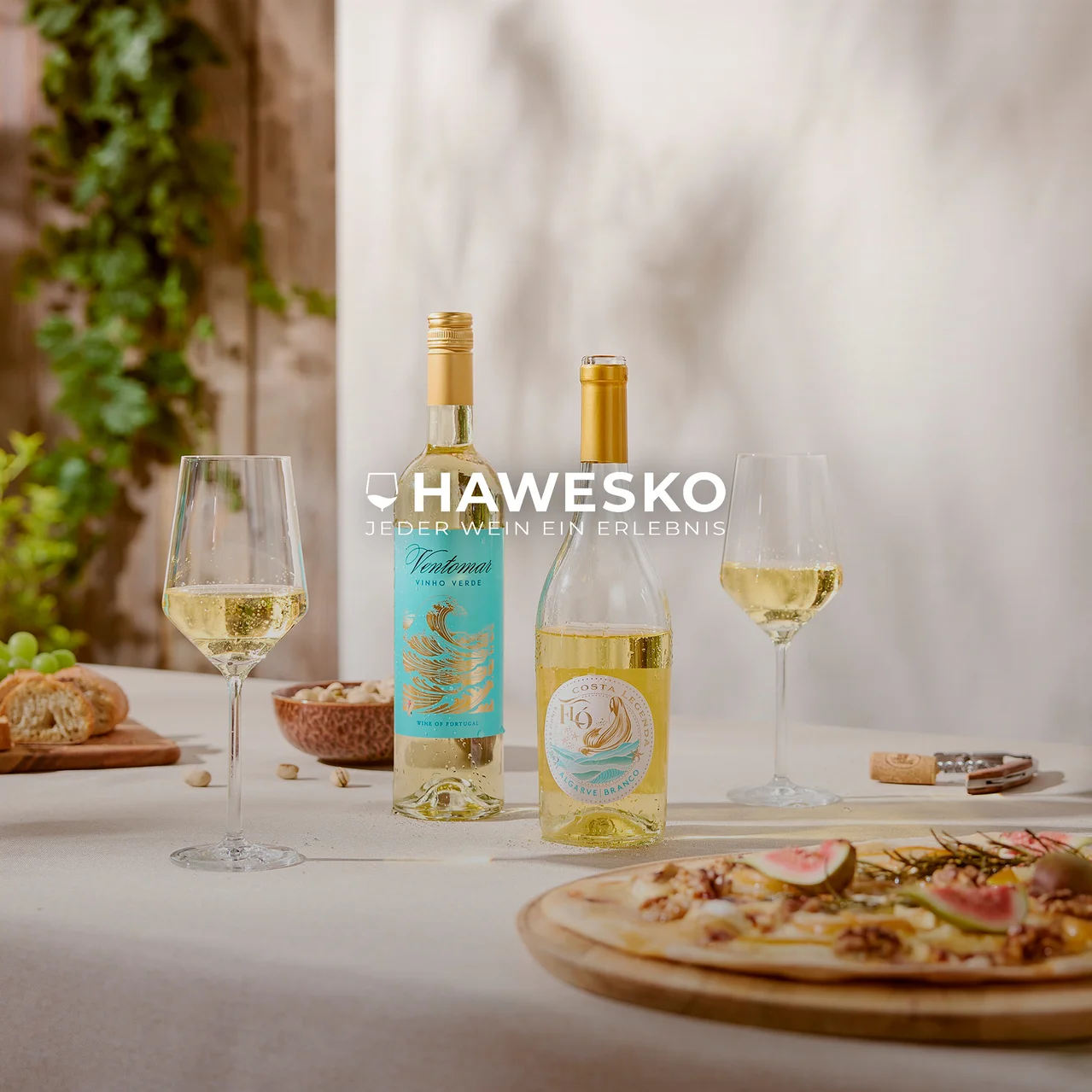 Zwei Flaschen Weißwein und zwei Gläser Weißwein auf einem Tisch mit Fladenbrot, Pistazien, Brot und Trauben. Im Hintergrund ist grünes Laub zu sehen. Text: HAWESKO JEDER WEIN EIN ERLEBNIS.