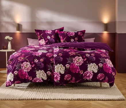 Ein Bett mit violetter Bettwäsche, die ein Muster aus rosa und weißen Blumen zeigt, steht in einem Schlafzimmer mit zweifarbigen Wänden und Holzboden. Auf einem Beistelltisch stehen eine Vase mit weißen Blumen und eine Kerze.