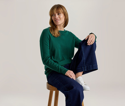 Frau in einem grünen Langarmshirt, weiten dunkelblauen Jeans und weißen Sneakern sitzt auf einem Holzhocker, blickt nach rechts und lächelt.