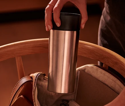 Eine Hand steckt einen silbernen Coffee-to-go-Becher in eine offene braune Ledertasche auf einem Holzstuhl.