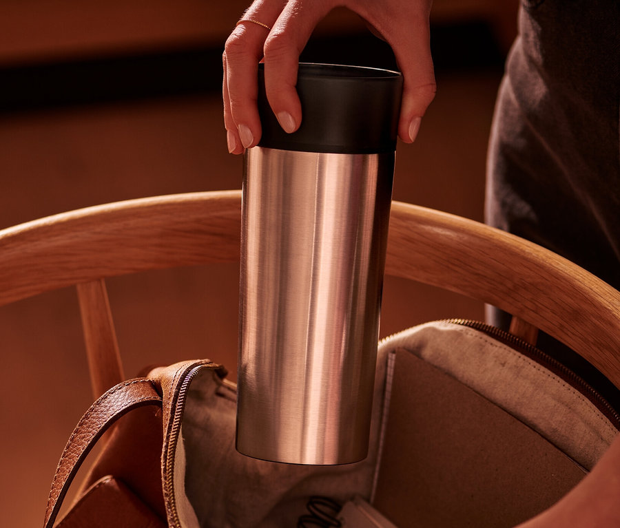 Eine Hand steckt einen silbernen Coffee-to-go-Becher in eine offene braune Ledertasche auf einem Holzstuhl.