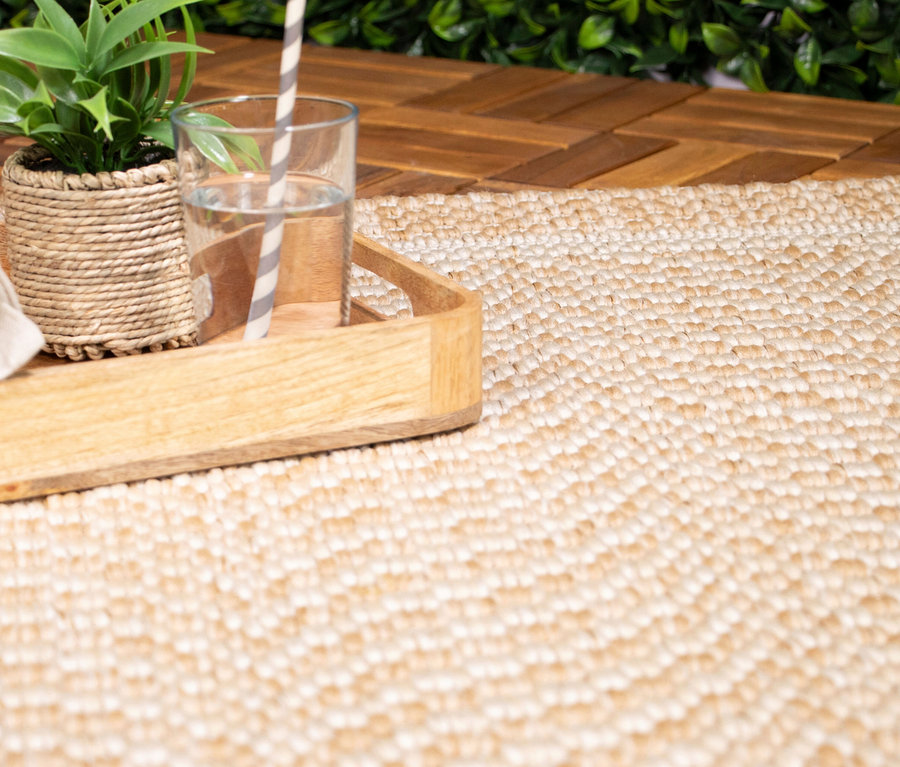 Nahaufnahme eines beigen Teppichs »Twine« mit Herringbone-Muster, auf dem ein Holztablett mit einer Pflanze und einem Wasserglas steht. Im Hintergrund sind Holzfliesen und grüne Pflanzen zu sehen.