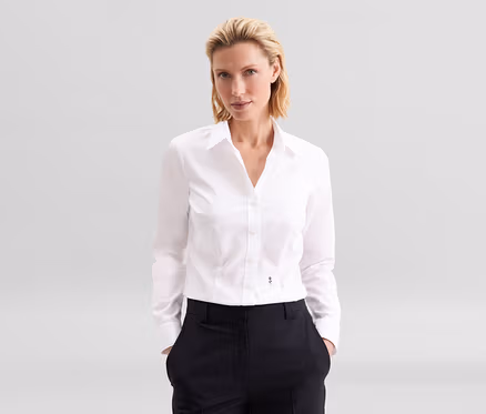 Blonde Frau in weißer Seidensticker Bluse Slim Fit non iron und dunkler Hose, Hände in den Taschen.