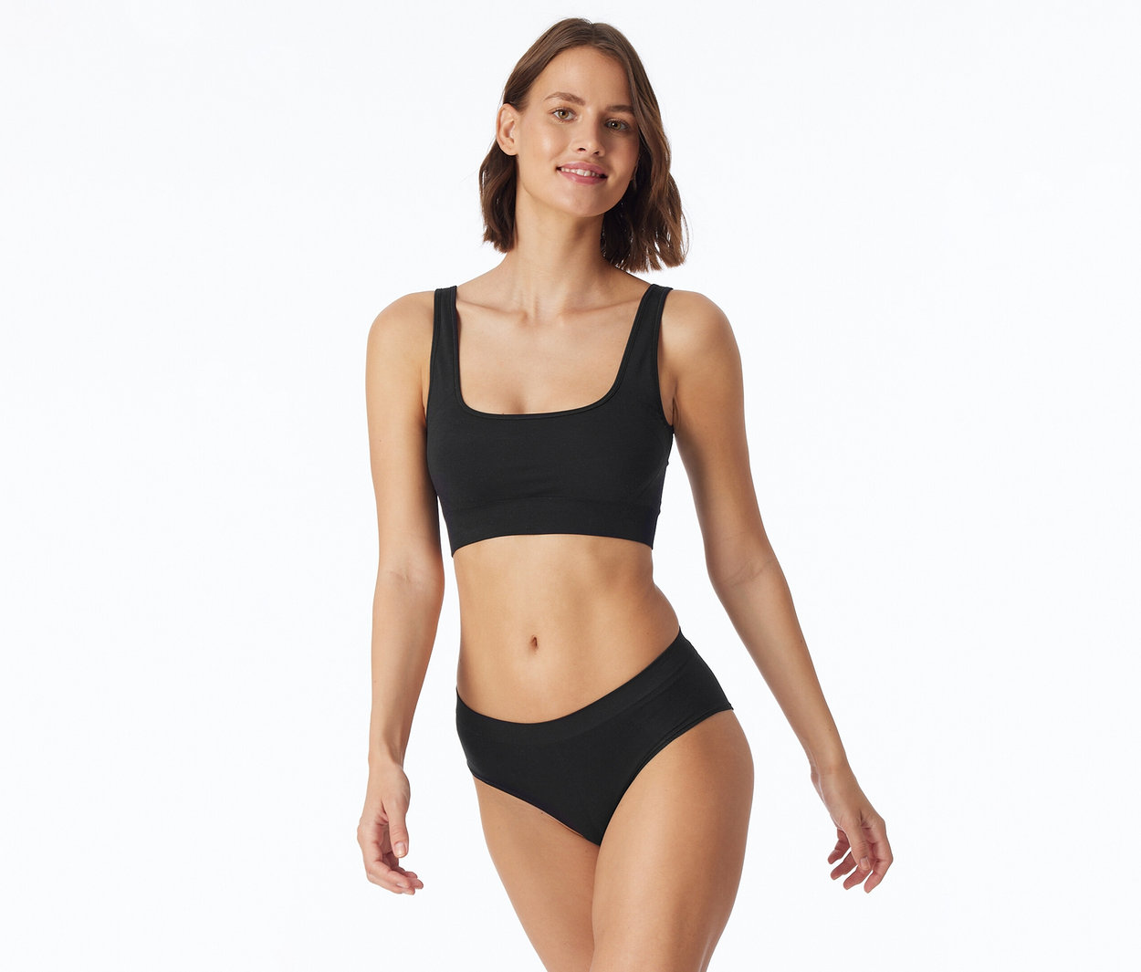 Junge Frau mit kurzen braunen Haaren, lächelnd, steht vor weißem Hintergrund und trägt einen schwarzen Bralette und schwarze Slips.