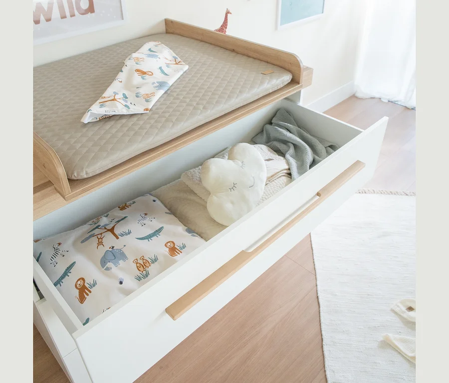 Babywickeltisch mit beige gesteppter Wickelauflage und einem geöffneten Schubfach, das Babybettwäsche mit Safarimotiven, ein herzförmiges Kissen und eine Decke enthält.