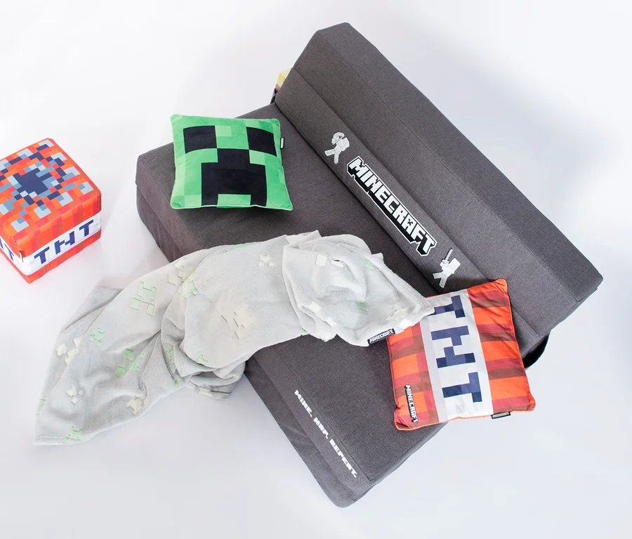 Ein graues Roba Minecraft Gaming Sofa ist auf einem weißen Hintergrund drapiert, zusammen mit einem TNT-Würfel, einem Creeper-Kissen, einem TNT-Kissen und einer grauen Decke.