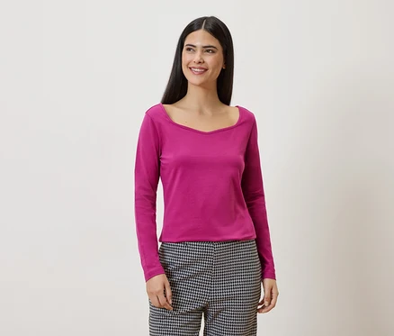 Frau mit langen, dunklen Haaren trägt ein pinkes Langarmshirt und eine schwarz-weiß karierte Hose vor einem weißen Hintergrund.