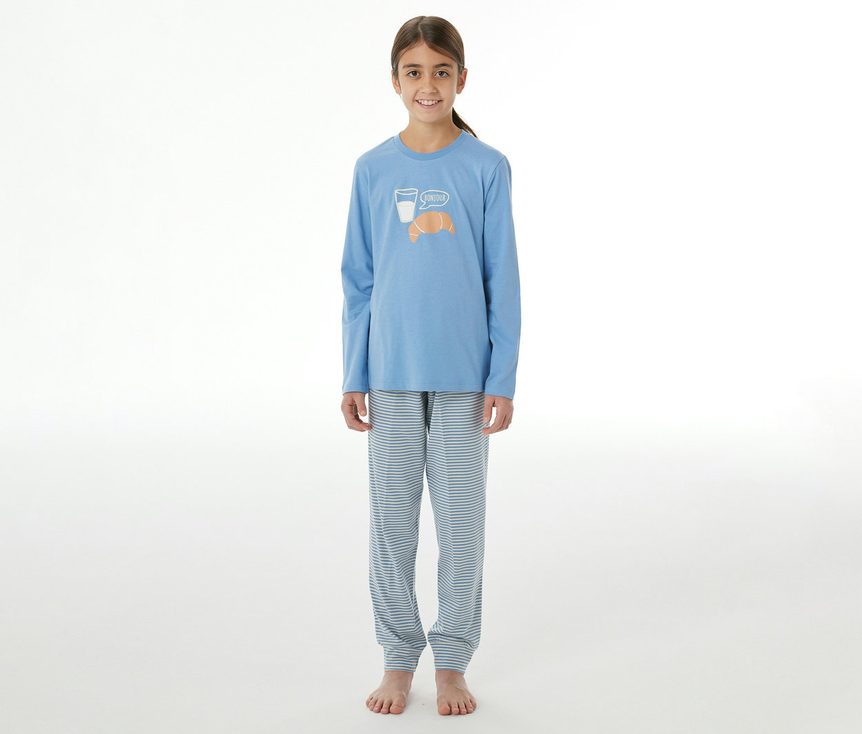 Junges Mädchen in einem blauen SCHIESSER Kinder-Pyjama mit Langarmshirt und gestreiften Hosen. Das Oberteil zeigt ein Glas Milch und ein Croissant mit „Bonjour“.