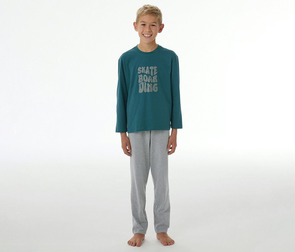 Ein lächelnder Junge steht barfuß und trägt einen SCHIESSER Kinder-Pyjama »Skate« mit einem dunkelgrünen Langarmshirt, auf dem "SKATE BOARDING" steht, und einer grauen Hose.