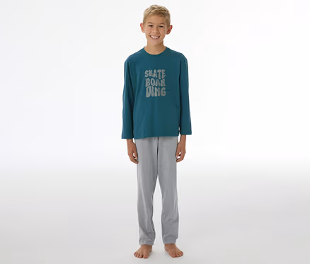 Ein lächelnder Junge steht barfuß und trägt einen SCHIESSER Kinder-Pyjama »Skate« mit einem dunkelgrünen Langarmshirt, auf dem "SKATE BOARDING" steht, und einer grauen Hose.