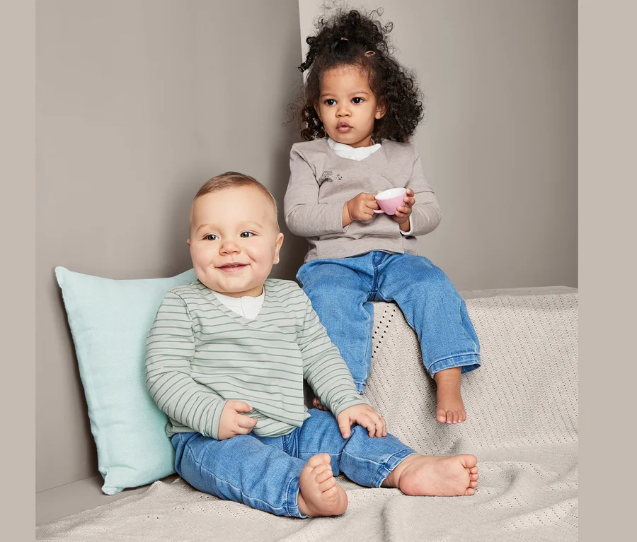 Ein Baby sitzt neben einem Kleinkind. Das Kleinkind hält eine rosa Tasse. Beide tragen Jeans und 2 Baumwoll-Jersey-Wickeljacken.