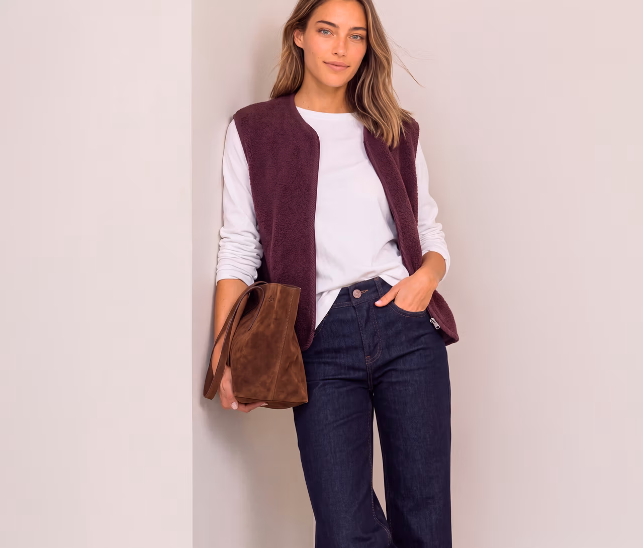 Frau in einer roten Street One Weste über einem weißen Langarmshirt und dunklen Jeans, die eine braune Handtasche hält und an einer Wand lehnt.