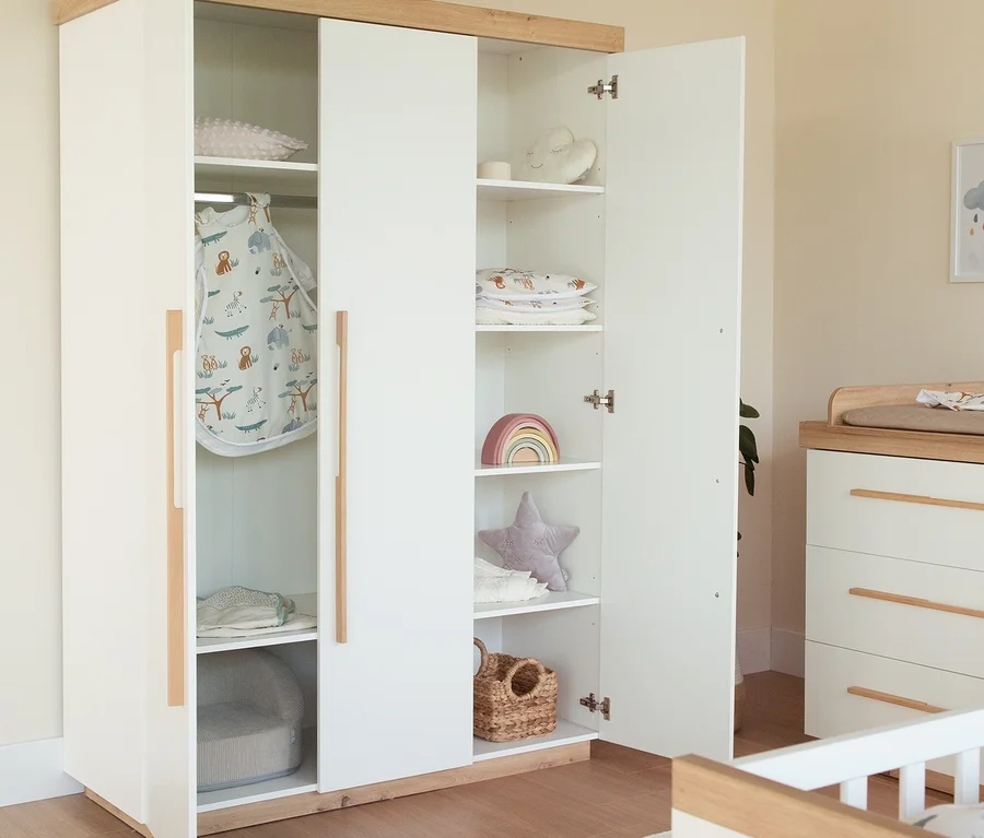 Ein offener weisser und hölzerner Roba Lumo Kleiderschrank, Teil eines 3-teiligen Kinderzimmer-Sets, gefüllt mit einem Schlafsack, gefalteter Kleidung und Spielzeug. Eine passende Wickelkommode und ein Gitterbett sind rechts teilweise sichtbar.