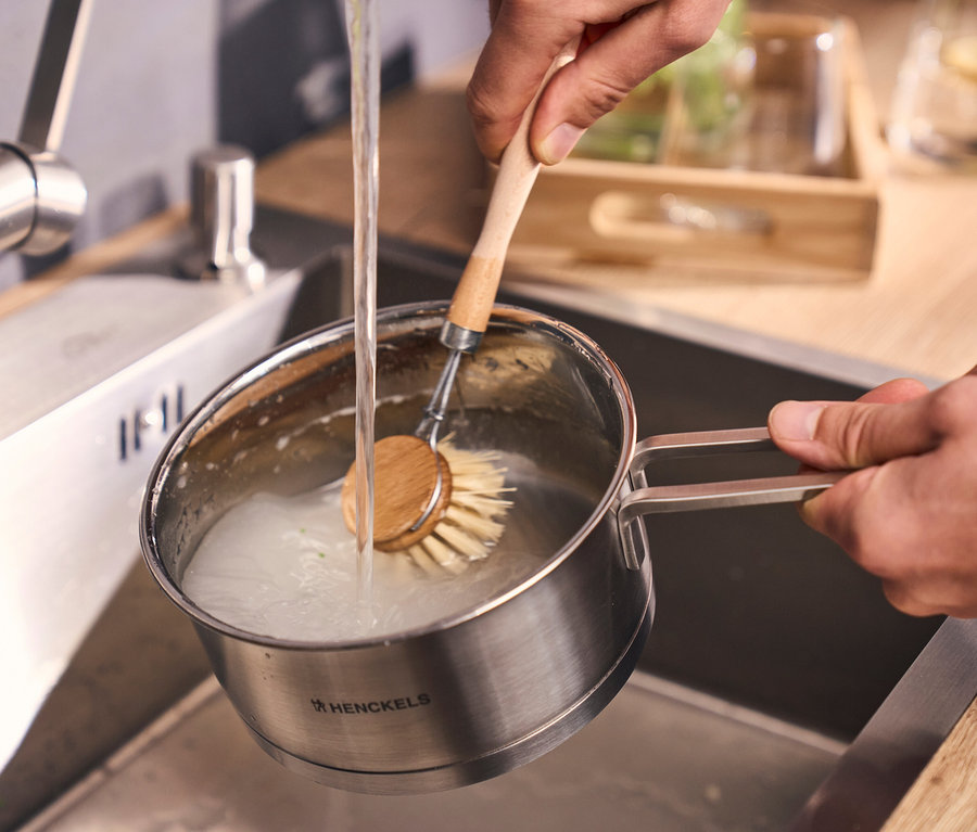 Hände spülen einen Topf aus dem Henckels by ZWILLING »Pulse« Kochgeschirrset mit einer Spülbürste unter fließendem Wasser in einem Spülbecken.
