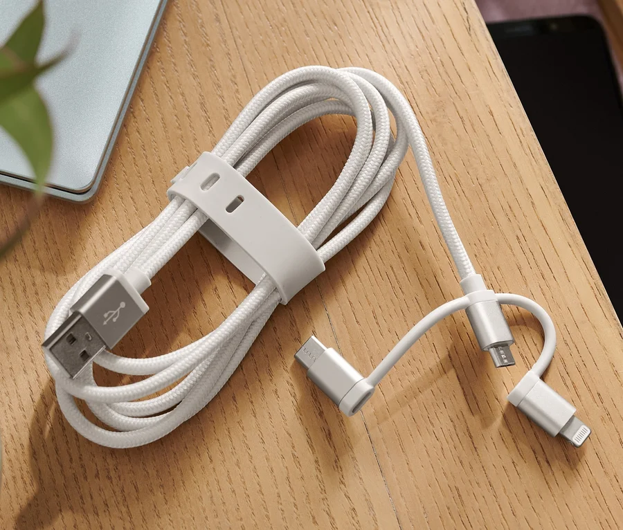 Ein graues 3-in-1-Ladekabel liegt auf einem Tisch neben einem Laptop und einer Vase mit einer Pflanze.