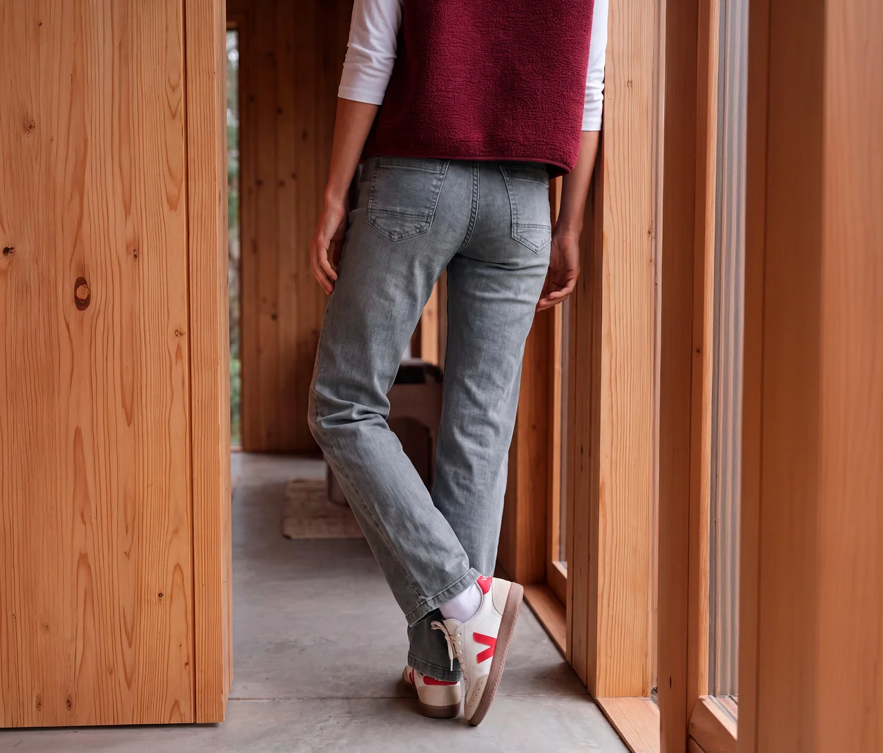 Person von hinten in hellgrauen CECIL Jeans, weißem Langarmshirt, bordeauxroter Weste und weißen Sneakers mit roten Akzenten, an Holzwand gelehnt.
