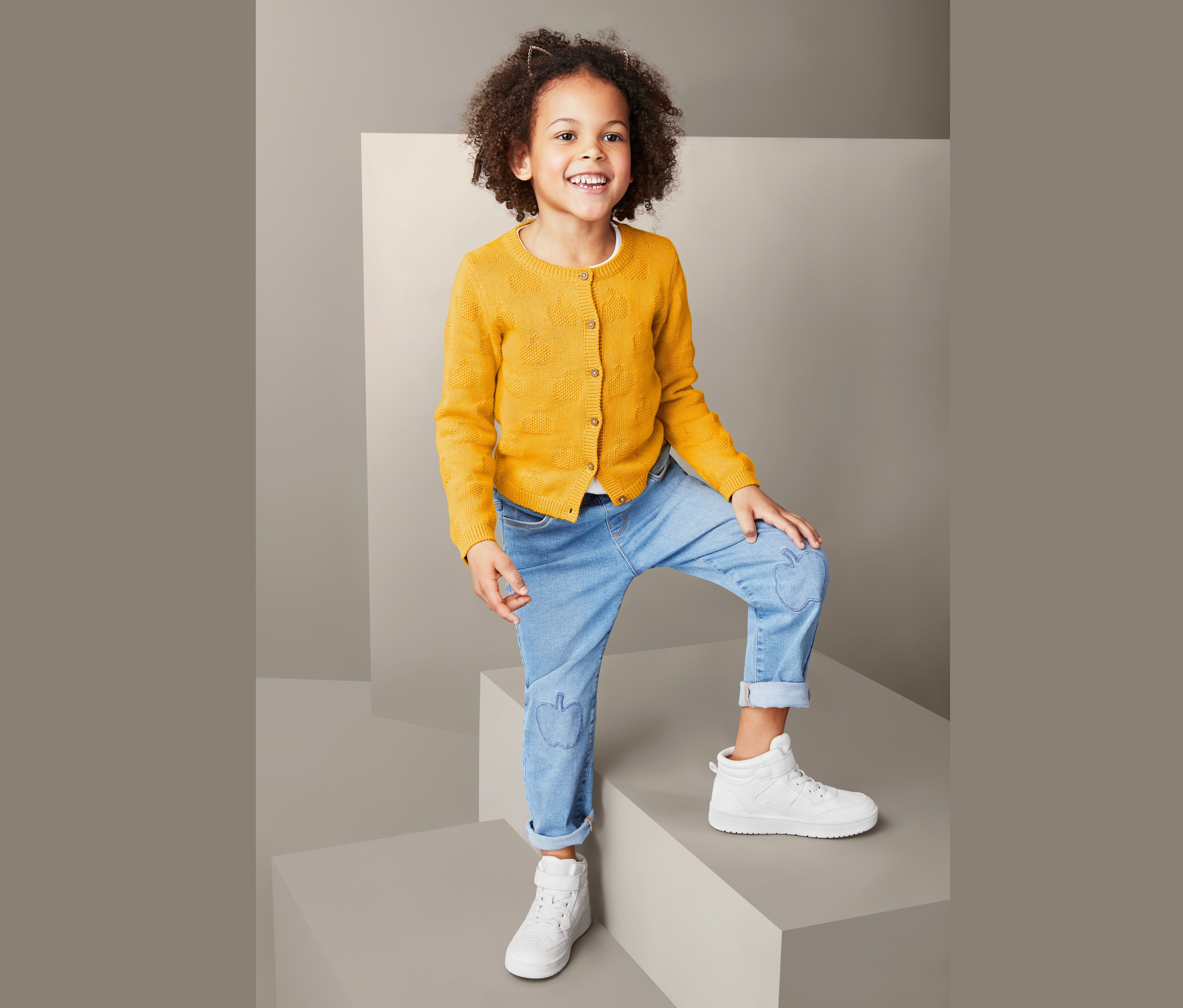 Mädchen mit lockigem Haar posiert in Strickjacke mit Apfelmotiv, Jeans und Turnschuhen.