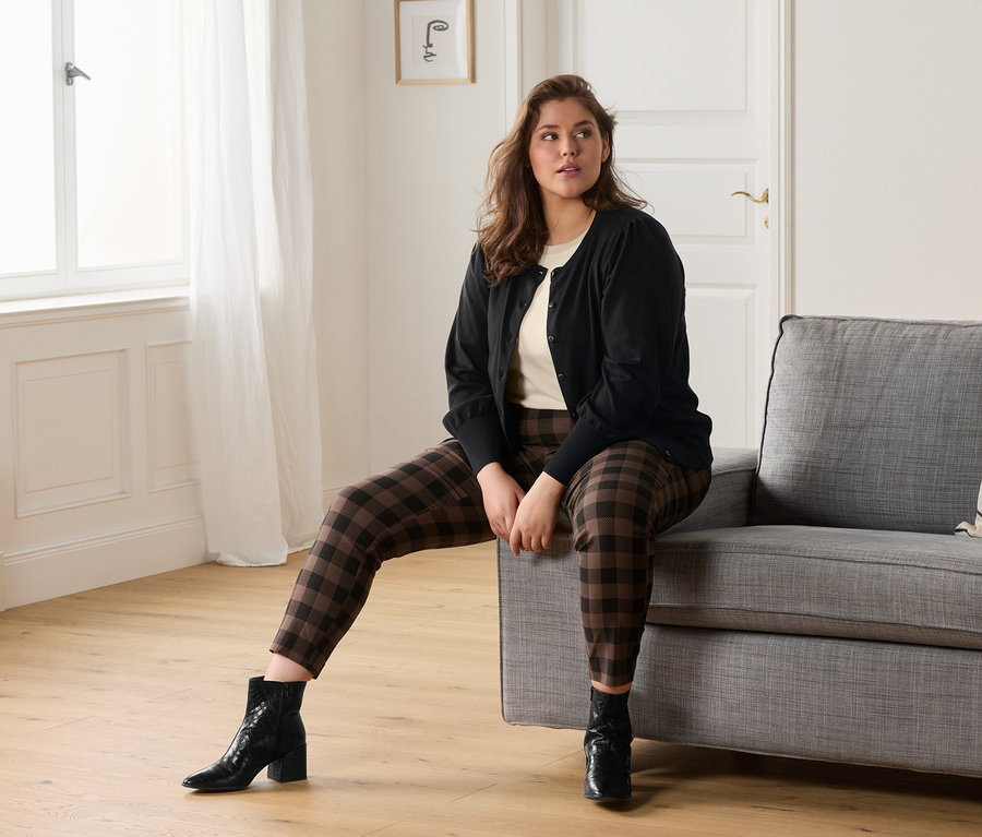 Frau sitzt auf einem grauen Sofa und trägt ein cremefarbenes T-Shirt, eine karierte Stretchhose und eine Feinstrickjacke mit Puffärmeln.