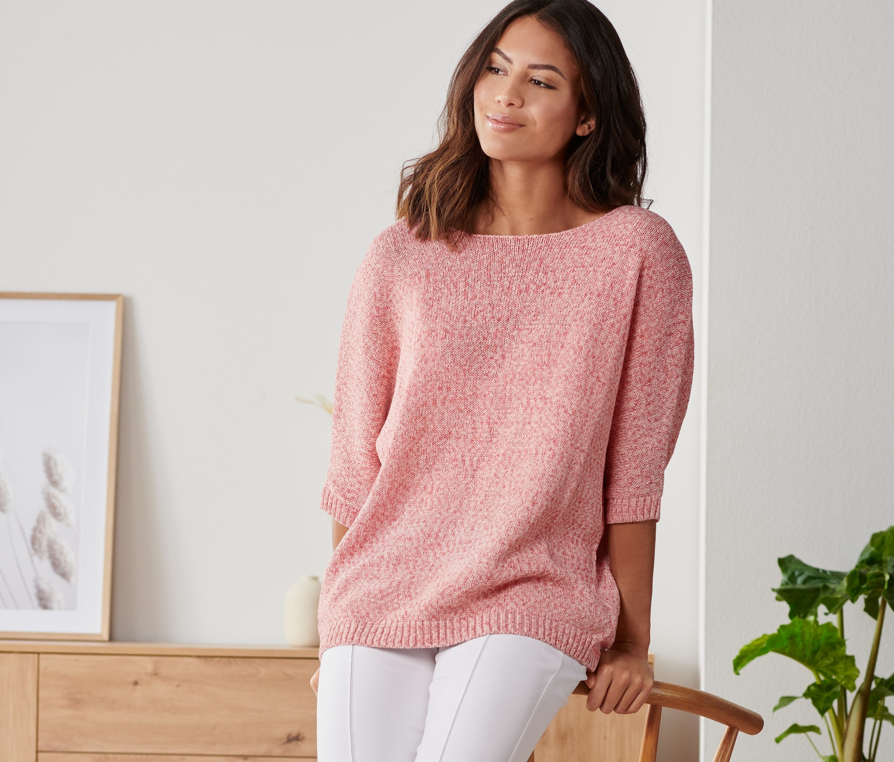 Eine Frau, die sich an einen Stuhl lehnt, trägt einen sommerlichen Strickpullover aus Bändchengarn, weiße Hosen und Turnschuhe.