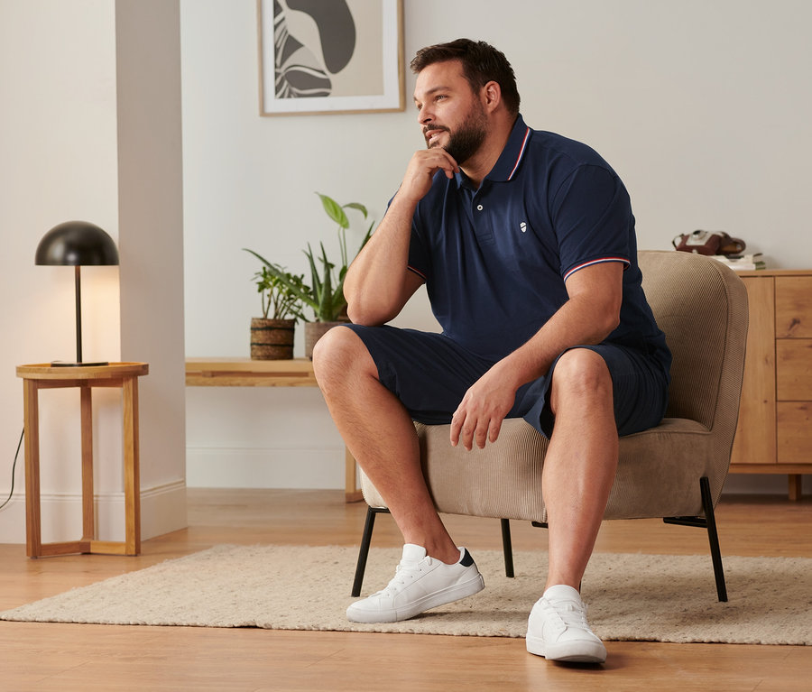 Mann sitzt auf einem Stuhl, trägt marineblaue Chino-Shorts, Sneaker und ein marineblaues Piqué-Poloshirt.