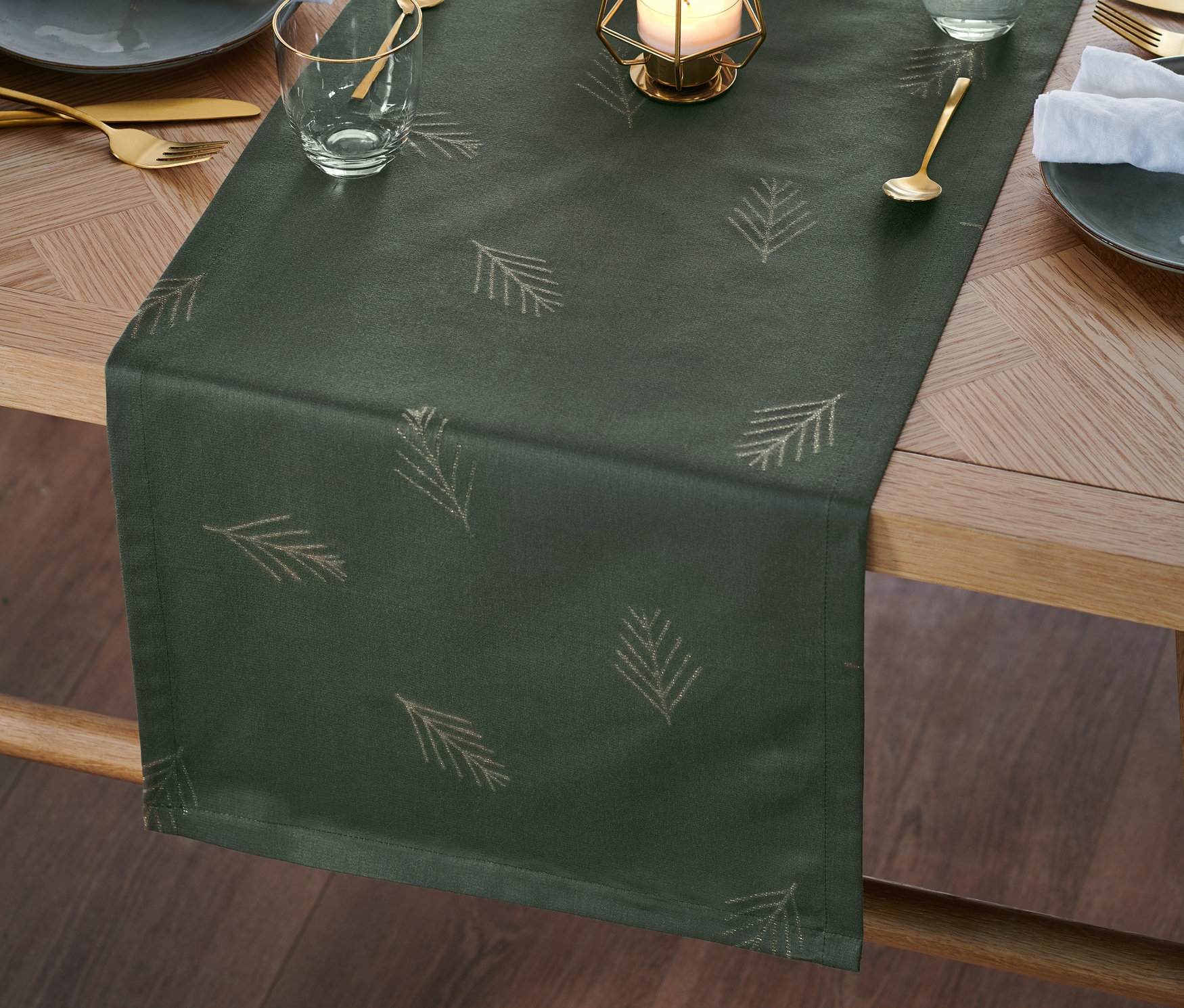 Auf einem Holztisch liegt ein grüner Jacquard-Tischläufer mit Nadelbaummotiv, dekoriert mit einer Kerze, Tannenzapfen und Geschirr.