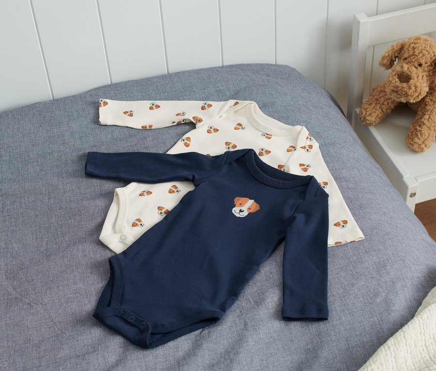 Zwei Babybodys liegen auf einem Bett. Ein Body ist dunkelblau mit einem Hundebild, der andere ist cremefarben mit einem Hundemuster.