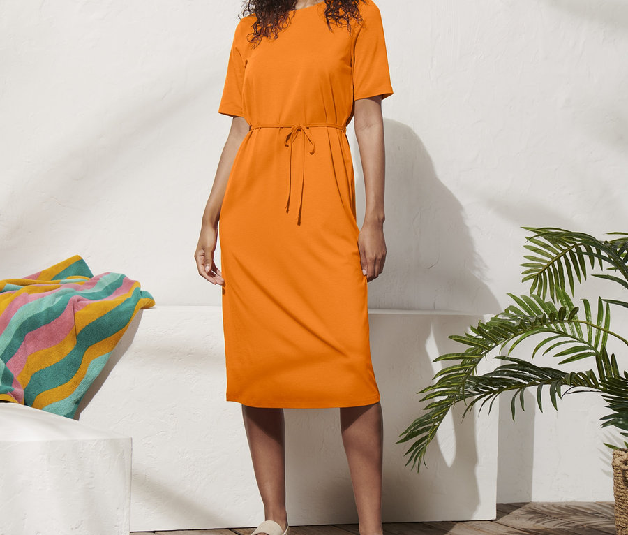 Frau steht in einem orangefarbenen Jerseykleid mit Taillengürtel und Hut.