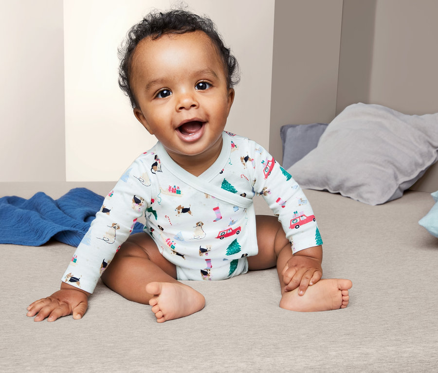 Lächelndes Baby sitzt auf einem Bett und trägt einen 3 Langarmbody mit Hunde- und Weihnachtsmuster.