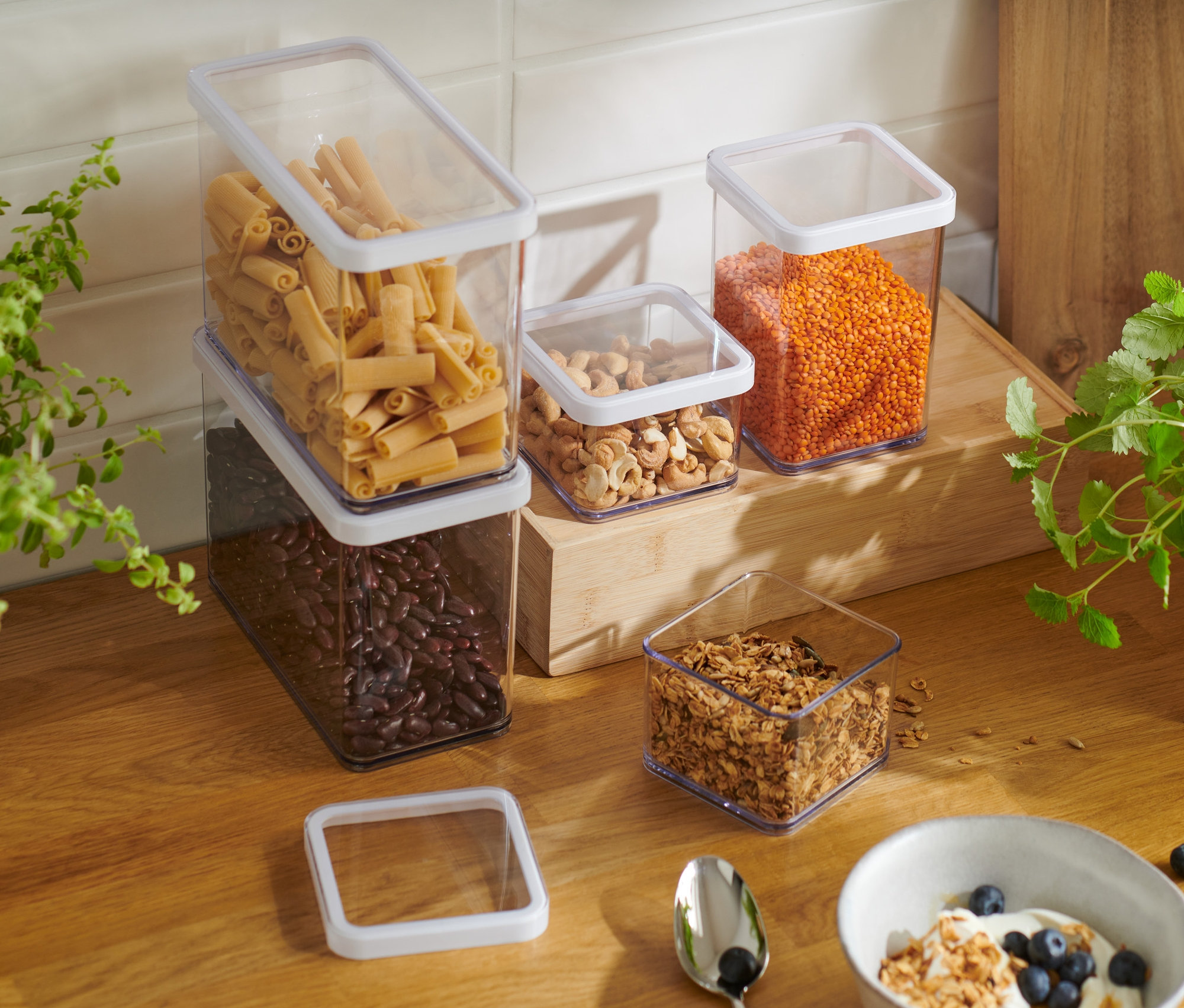 Fünf unterschiedlich große Vorratsdosen, gefüllt mit Bohnen, Nudeln, Nüssen und Linsen, stehen auf einer hölzernen Küchentheke neben einer Schüssel mit Joghurt und Müsli.