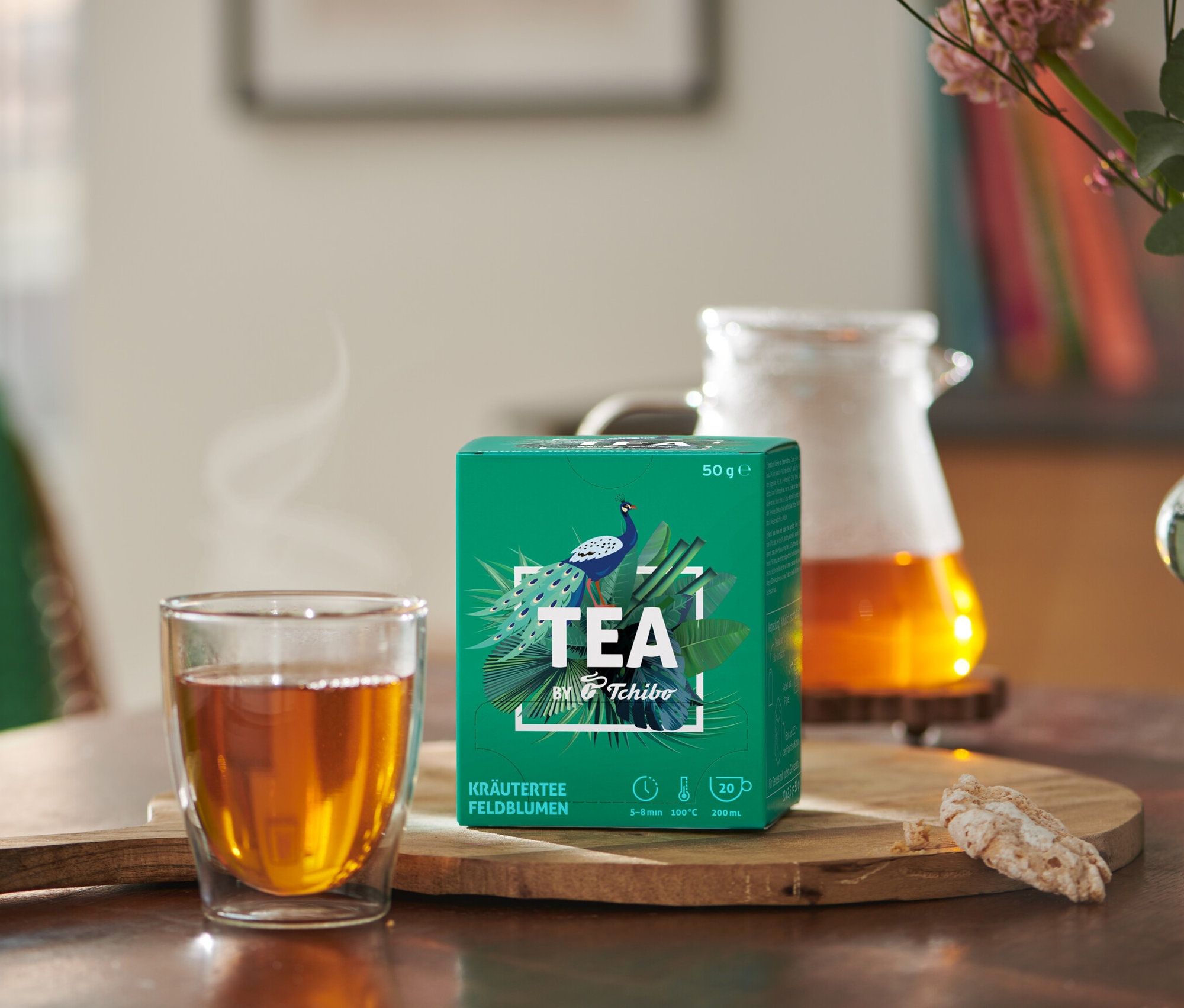 Auf einem Tisch stehen eine Tasse Tee und eine Packung TEA by Tchibo Kräutertee Feldblumen Teebeutel.