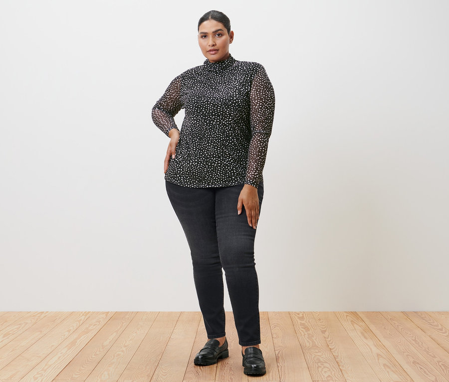 Frau posiert in einem gepunkteten Mesh-Rollkragenshirt, schwarzen Jeans und Schuhen.