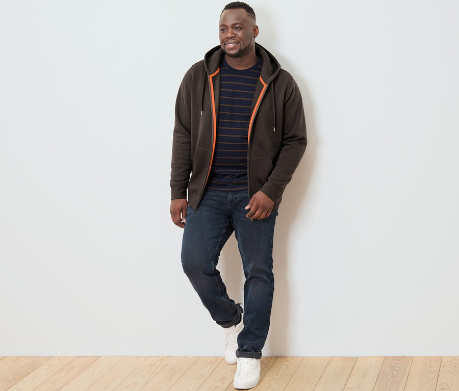 Mann posiert auf Holzboden vor einer weißen Wand und trägt eine braune Kapuzenjacke.