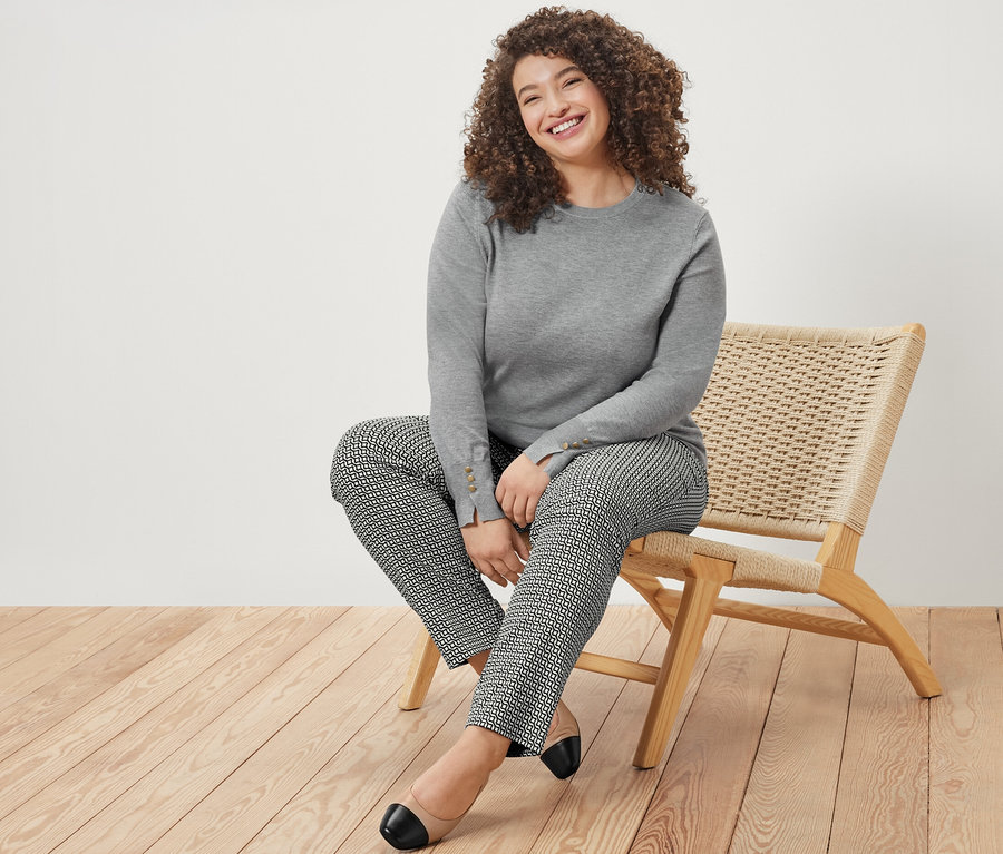 Eine lächelnde Frau sitzt auf einem Stuhl und trägt einen grau melierten Feinstrickpullover und eine bedruckte Stretchhose in Navy.