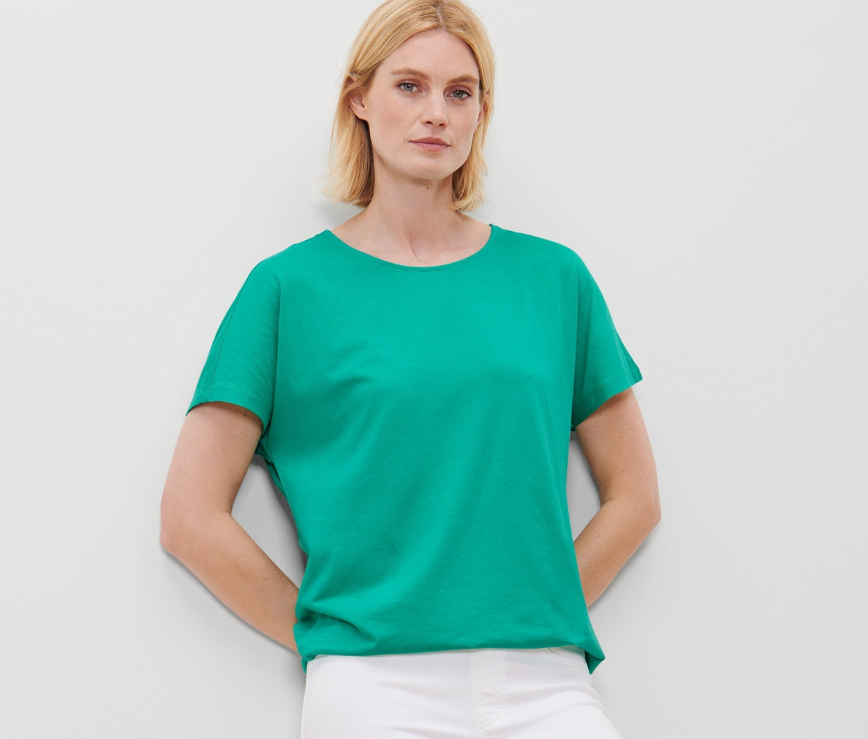 Blonde Frau posiert in grünem Blusenshirt und weißen Hosen.