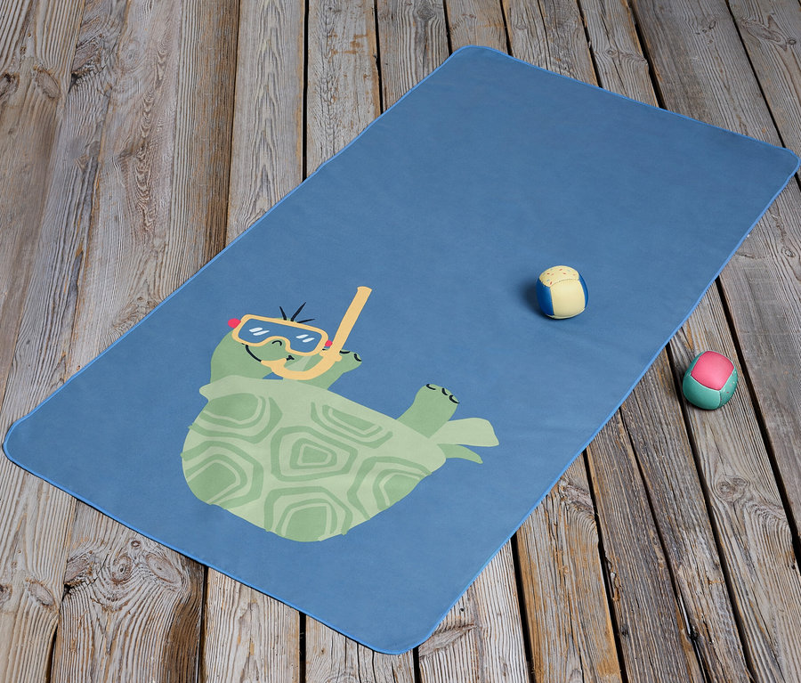 Blaues Kinder-Microfaserhandtuch mit Comic-Schildkröte mit Brille und Schnorchel liegt auf einem Holzboden. Neben dem Handtuch liegen zwei kleine Bälle.