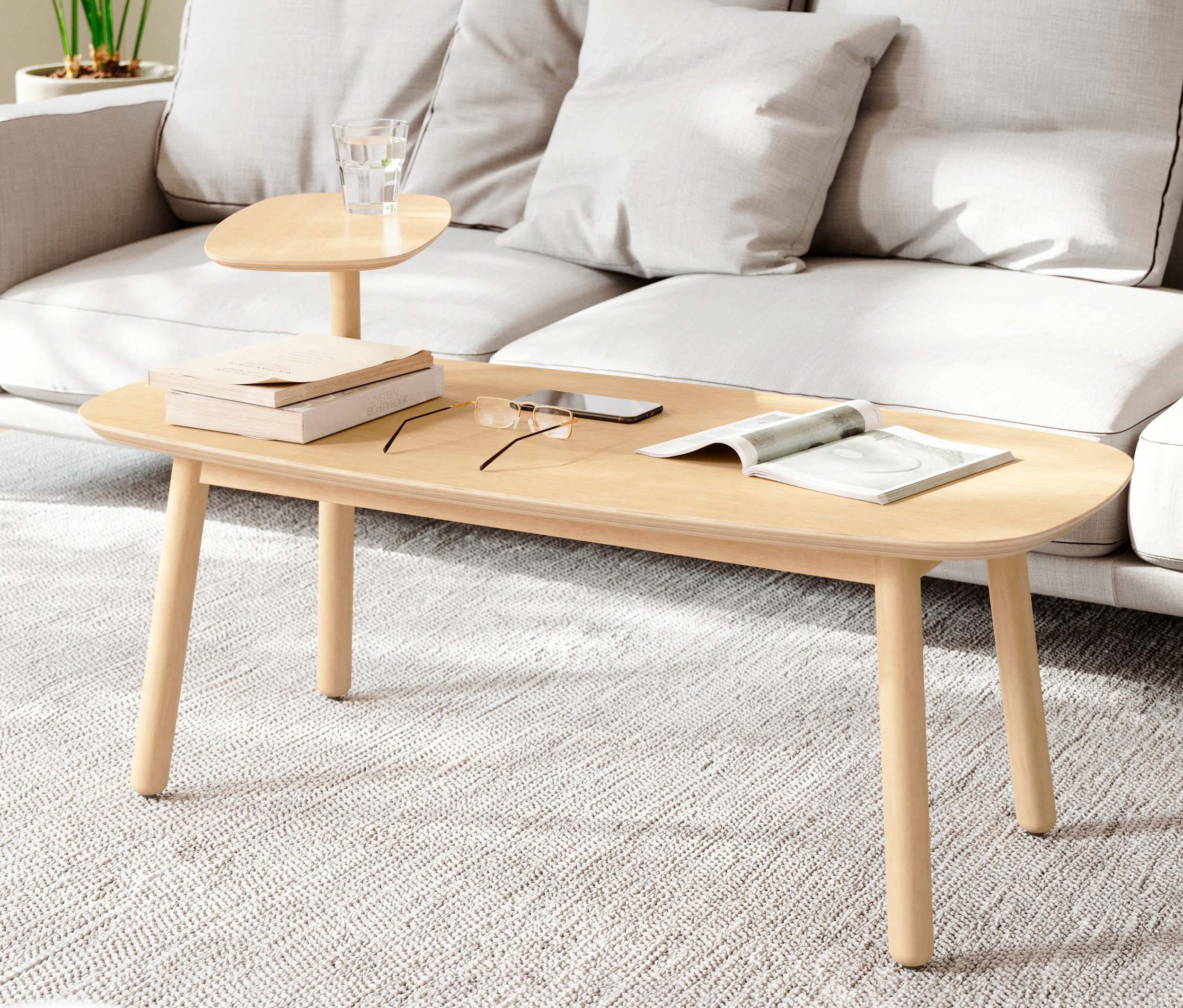 Beiger Couchtisch mit Büchern, Brille, Telefon und Zeitschrift vor einem grauen Sofa.