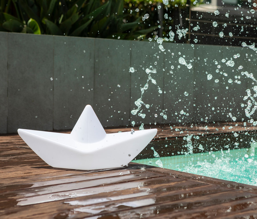 Die weiße Goodnight Light LED-Leuchte »Boot« liegt auf einem Holzboden neben einem Pool mit spritzendem Wasser.