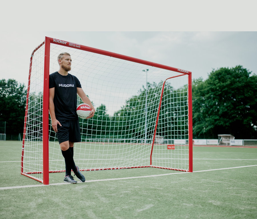 Mann in schwarzer Sportbekleidung steht an einem rot-weißen Fußballtor und hält einen Fußball.