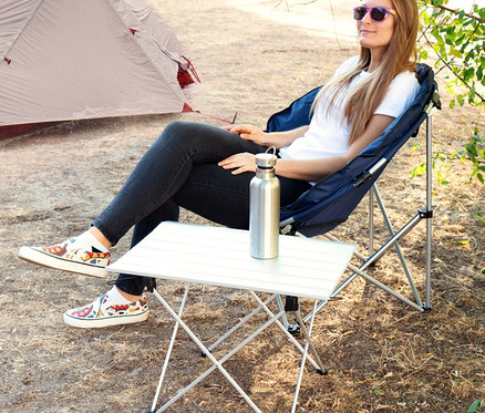 Frau sitzt auf Campingstuhl neben faltbarem Navaris Campingtisch und Edelstahlwasserflasche.