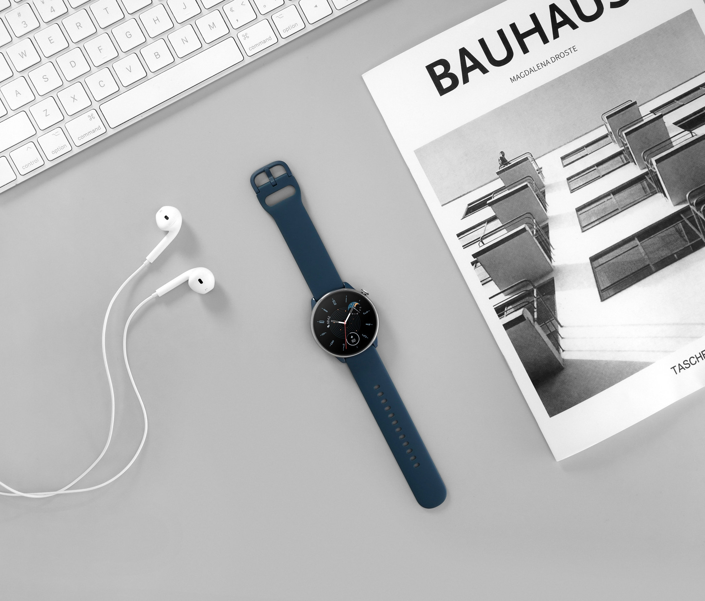 Auf einem grauen Hintergrund liegen eine weiße Tastatur, weiße Kopfhörer, eine blaue Amazfit GTR Mini Smartwatch und ein Bauhaus-Buch.