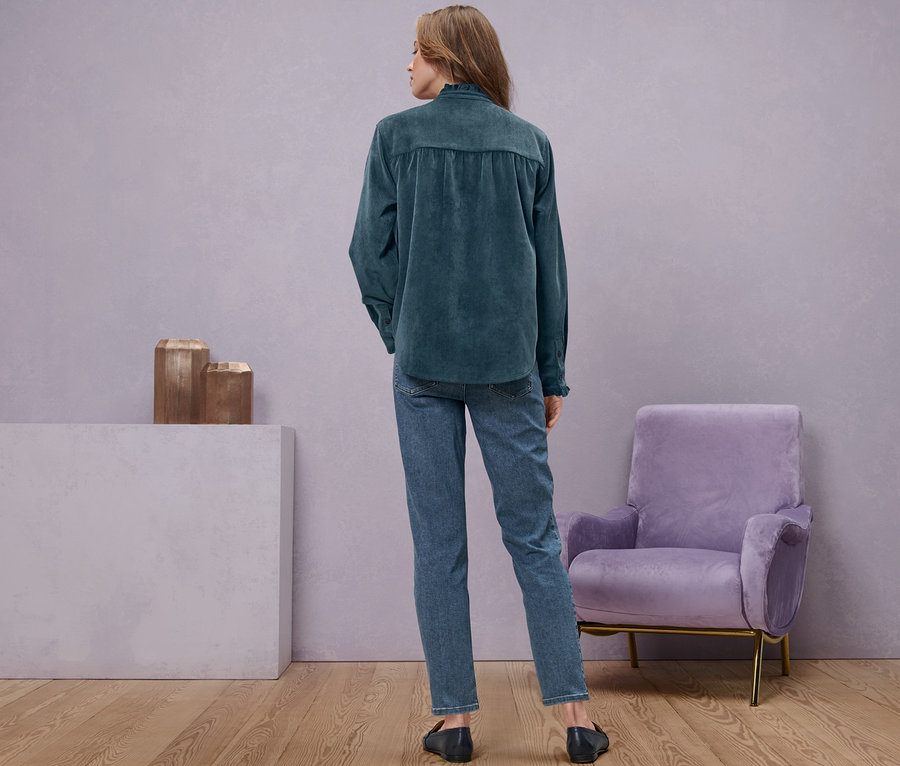 Eine Frau steht mit dem Rücken zur Kamera und trägt Jeans und eine Cordbluse mit Rüschendetail.
