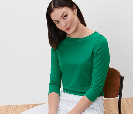 Frau sitzt auf einem Stuhl und trägt ein grünes Shirt mit 3/4-Arm und weiße Hosen.